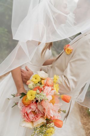 A spring wedding bouquet featuring coral peonies, yellow ranunculus, orange tulips and delicate white veil in the background.