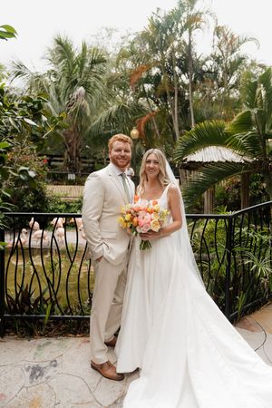 A wedding couple poses in front of lush tropical palm trees and greenery at an outdoor garden venue.