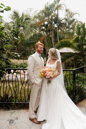 A couple in elegant wedding attire pose together on a tropical garden pathway.