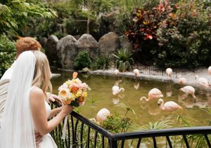 A bride admires flamingos in a pond while holding a peach and coral bouquet.