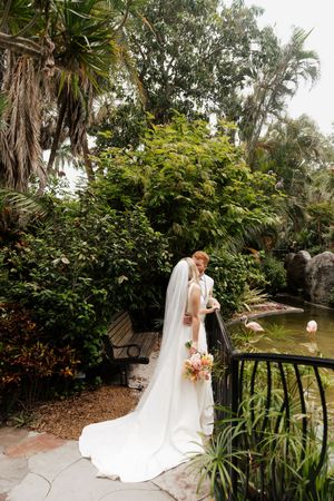 A wedding couple poses in a lush garden setting with flamingos visible in the background.