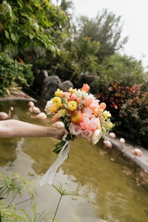 A delicate bouquet of pink, yellow and coral flowers with white ribbon held over a garden pond.