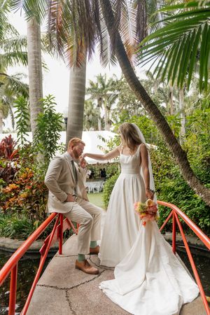 Romantic wedding couple poses on red bridge surrounded by tropical palm trees and lush gardens.
