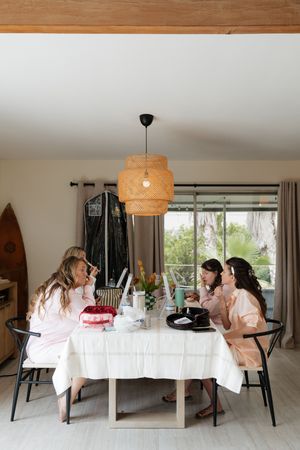 A group gathers around a white dining table with a rattan pendant light hanging above in a modern coastal interior.