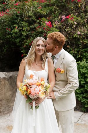 A romantic wedding portrait in a garden with blooming red flowers and a peach and coral bouquet.