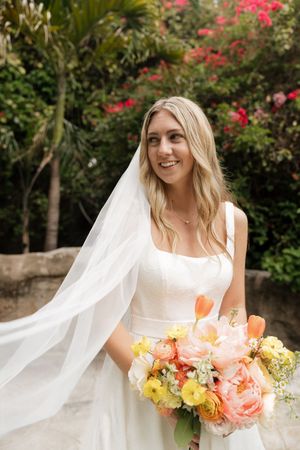 A bride in a flowing white dress holds a vibrant peach and yellow bouquet against a floral backdrop.