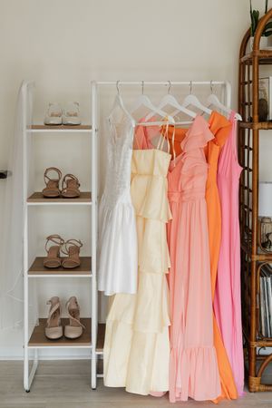 A collection of pastel and white dresses hanging on a white shelf unit with brown shoes displayed on the side.
