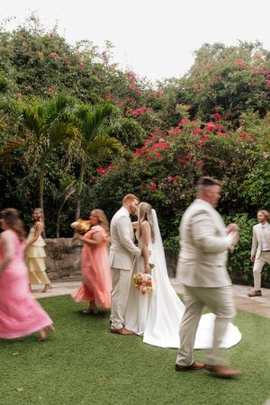 Wedding party dances on green lawn with pink flowering bushes in background creating joyful movement.