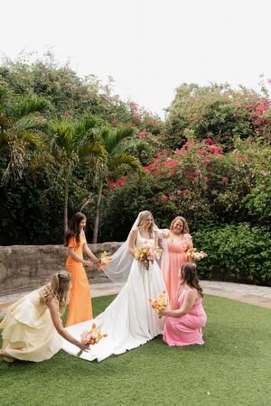 Bridesmaids in pastel dresses help arrange bride's gown on green lawn.
