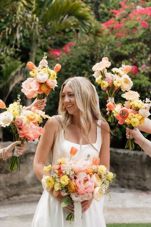 Bridesmaids in white dresses holding vibrant coral and peach floral bouquets against tropical garden backdrop.