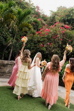 Wedding party in colorful dresses dancing and celebrating in a tropical garden setting with pink flowers.