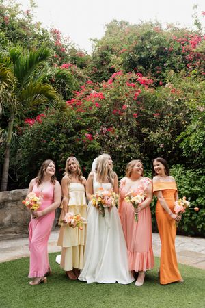 Bridesmaids in pastel dresses ranging from pink to orange pose together in a tropical garden setting.