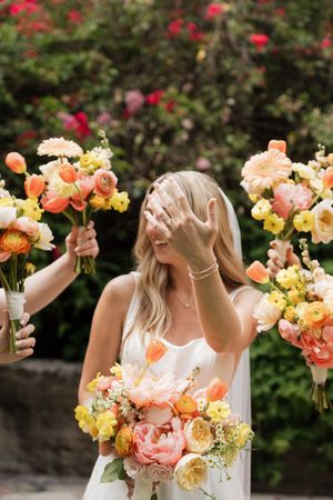 Bridesmaids hold beautiful peach and yellow floral bouquets against a backdrop of red flowering bushes.