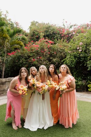 Bridal party posing together in sunset colors of pink, orange and cream dresses against flowering bushes.