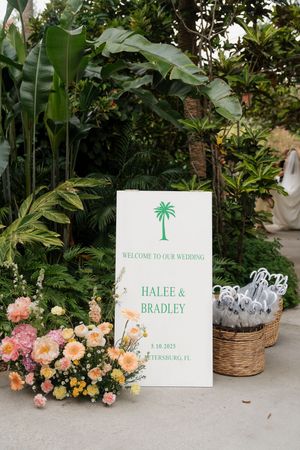 Wedding welcome signs with palm tree motifs displayed among tropical greenery and coral-colored flowers.