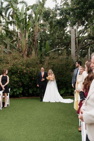 Wedding ceremony taking place on a manicured lawn surrounded by tropical palm trees and greenery.