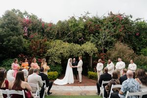 A panoramic view of an outdoor wedding ceremony with guests seated in white chairs surrounded by lush gardens and trees.