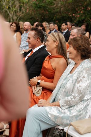 Wedding guests seated outdoors watching ceremony with a woman in orange dress in focus.