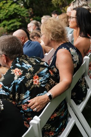 Wedding guests seated in white chairs wearing floral dresses at outdoor ceremony.