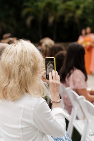 Wedding guests seated in white chairs view a ceremony through raised phones and cameras.