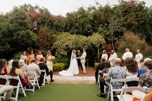Outdoor garden wedding ceremony with rows of white chairs surrounded by blooming trees and landscaping.