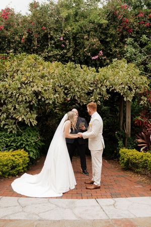 A wedding ceremony takes place in a lush garden setting with red flowers and brick pathways.