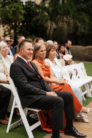 Wedding ceremony guests seated in white chairs on grass with formal attire and palm trees in background.