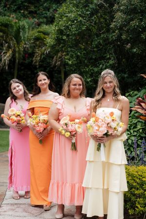 Bridesmaids in coordinating peach, orange and cream colored maxi dresses hold matching floral bouquets in a garden setting.