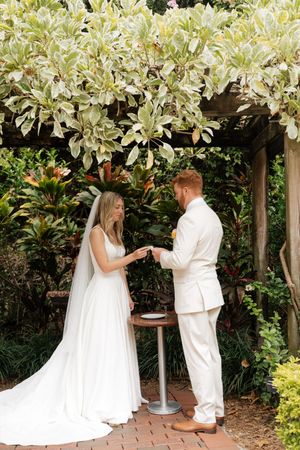 A couple in white attire dancing together under a garden pergola covered in flowering vines.