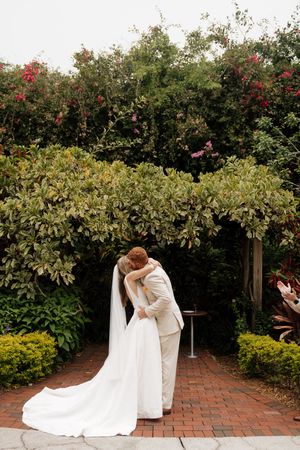 A romantic wedding couple embraces on a brick path surrounded by lush garden hedges with red flowers.