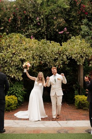A couple celebrates joyfully in a garden setting with brick pathways and flowering bushes.