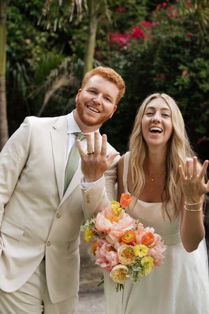 Wedding couple in white attire laughing and celebrating with bright coral and yellow floral bouquet.