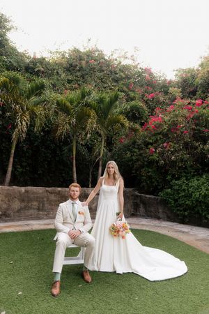 A wedding couple sits on a bench in a lush tropical garden setting with pink flowers and palm trees.