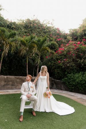 A wedding couple relaxes on grass surrounded by tropical foliage and pink bougainvillea flowers.
