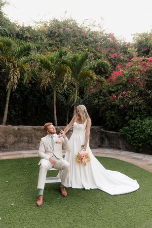 Wedding couple posing playfully on grass with white dress trailing against tropical garden backdrop.