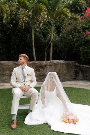 Bride and groom in elegant white attire seated on grass with tropical garden backdrop.