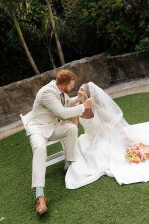 Wedding couple in white attire pose playfully on garden lawn.