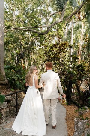 Newlyweds walk hand in hand down a tropical garden path surrounded by palm trees.