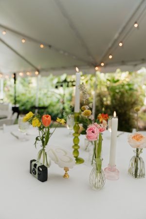 Simple wedding table decorations with small vases of colorful flowers under a white tent with string lights.