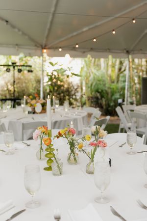 Wedding table settings with delicate floral centerpieces in bud vases under a tent with string lights.