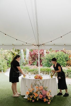 Event staff arrange floral decorations on a table under a white tent.