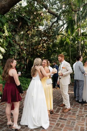 Wedding party mingles on a brick courtyard under lush tropical foliage.