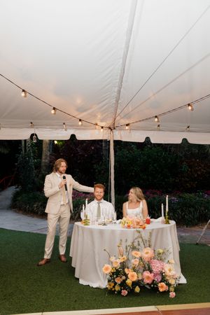 Wedding reception setup under white tent with elegant table settings and colorful floral arrangements.
