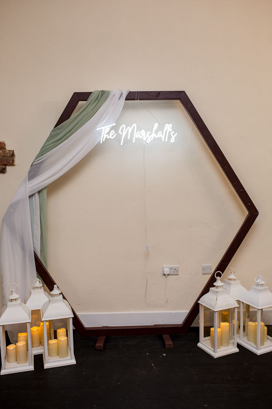 Decorative hexagonal wedding sign with white draping and lantern decorations against a neutral wall.