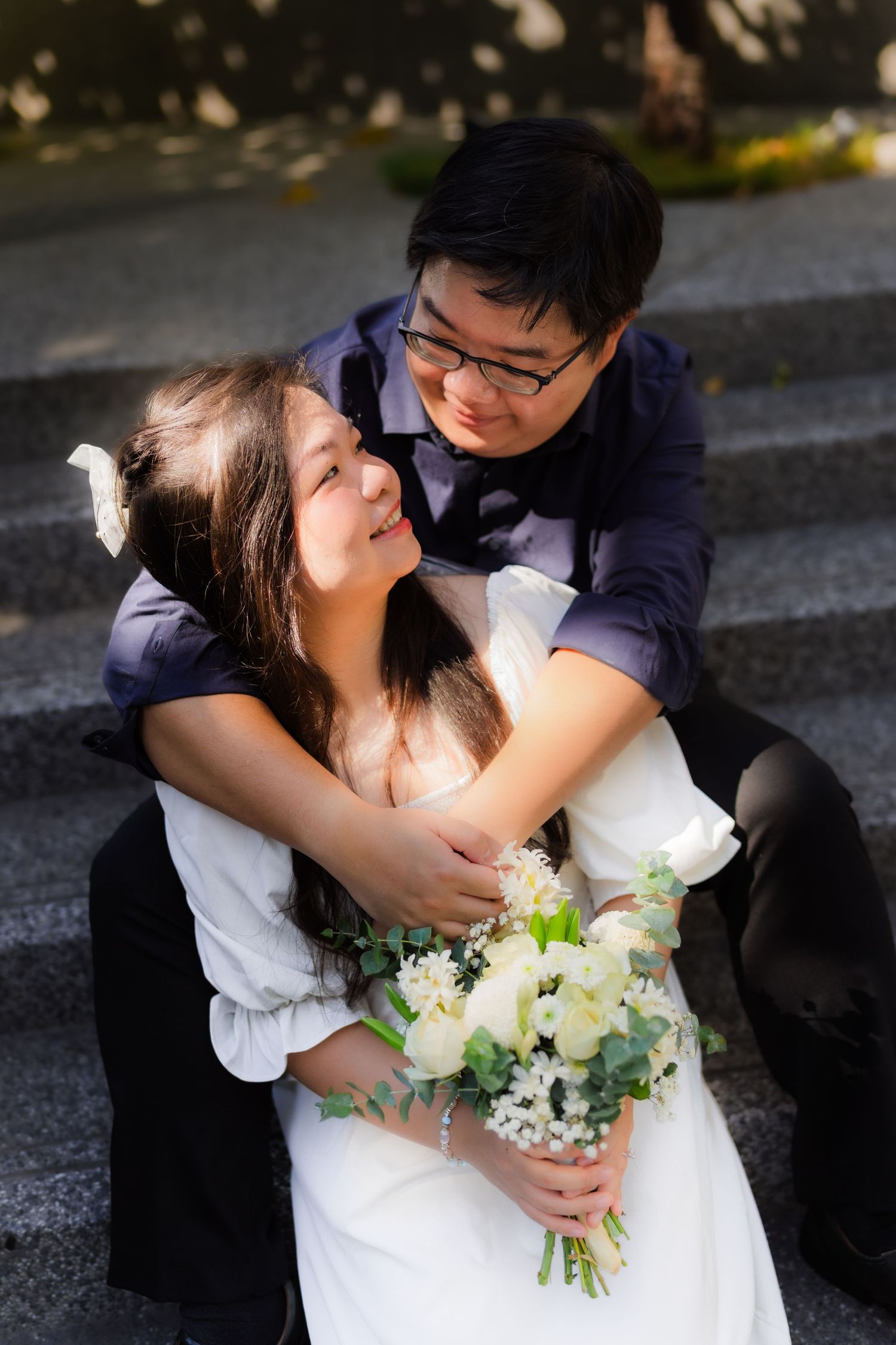 A romantic wedding couple embraces on stone steps while holding a bouquet of white and green flowers.
