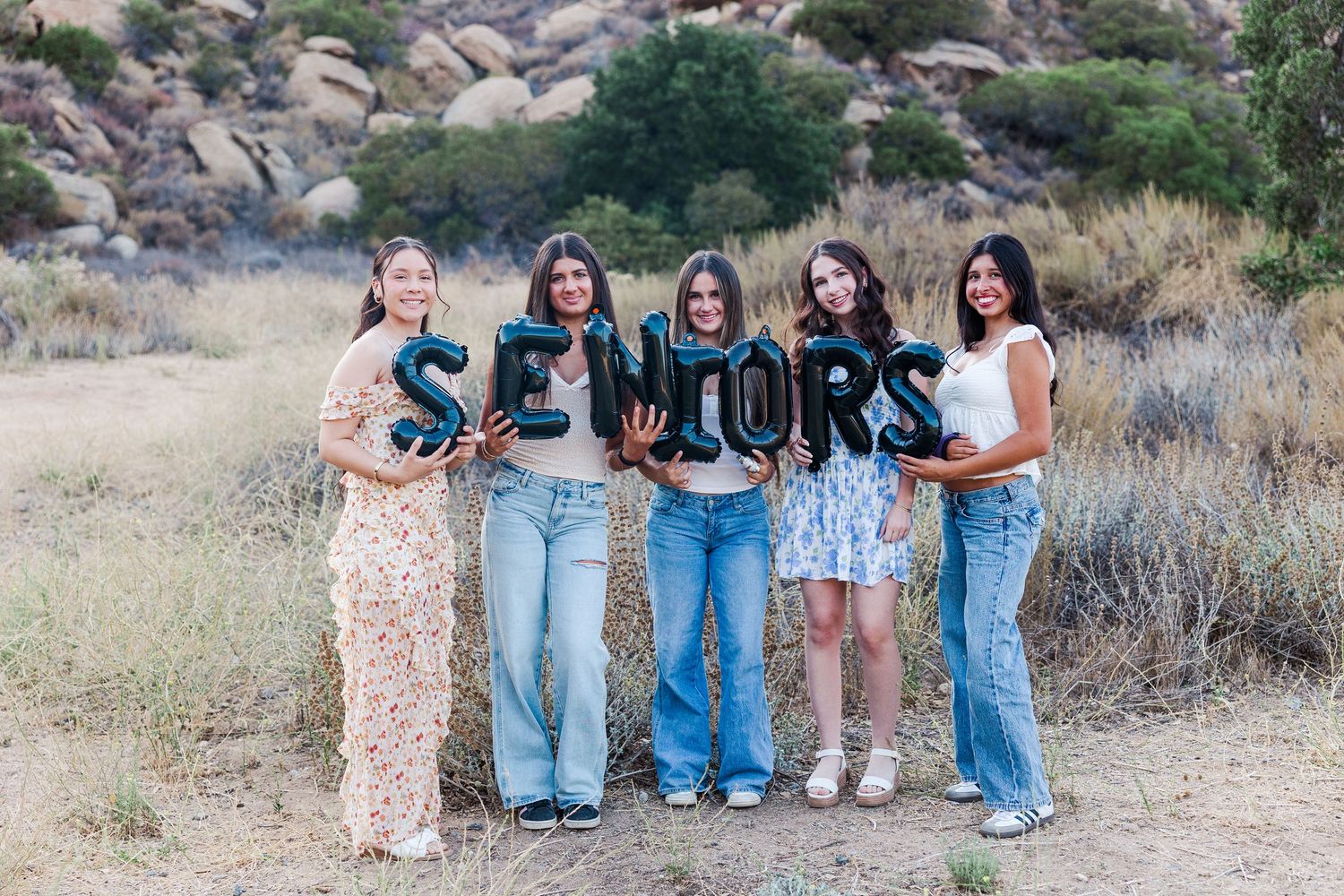 Group of friends pose with SENIORS letters outdoors in a desert landscape with rocks and brush in background.
