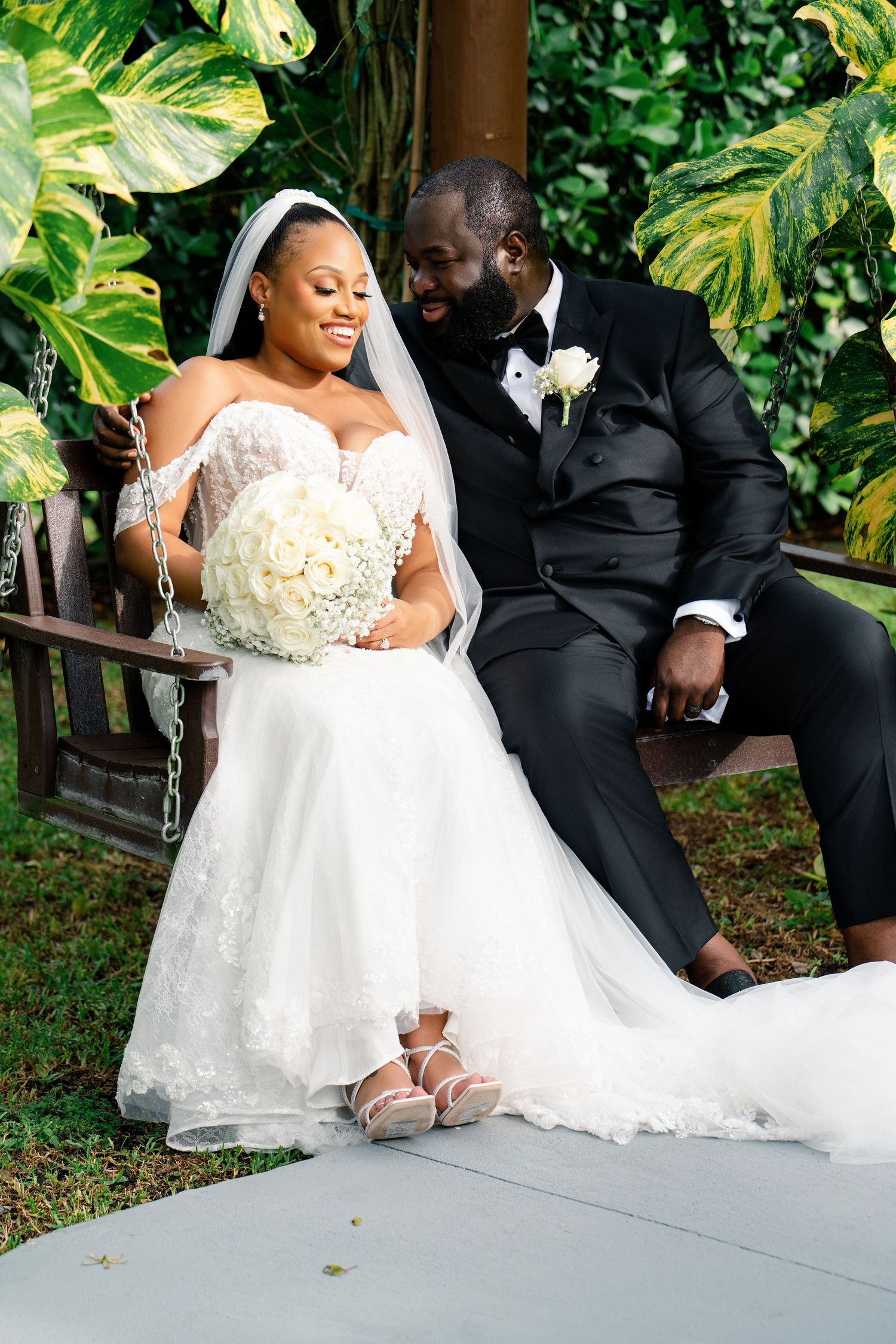A romantic wedding couple shares intimate moments while sitting on a garden swing surrounded by lush greenery.