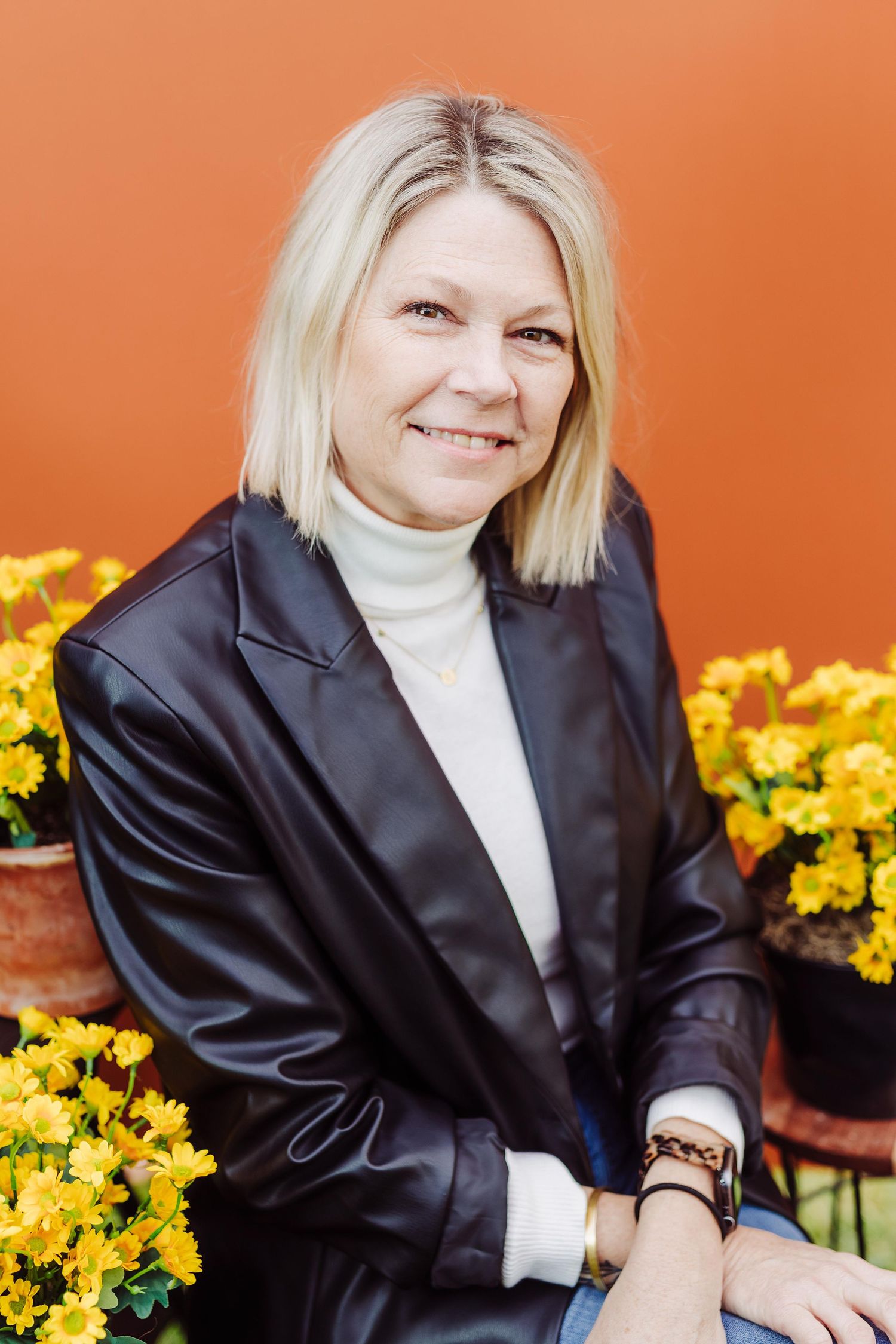 Professional portrait against orange wall with yellow flowers, wearing black leather jacket and white turtleneck sweater.