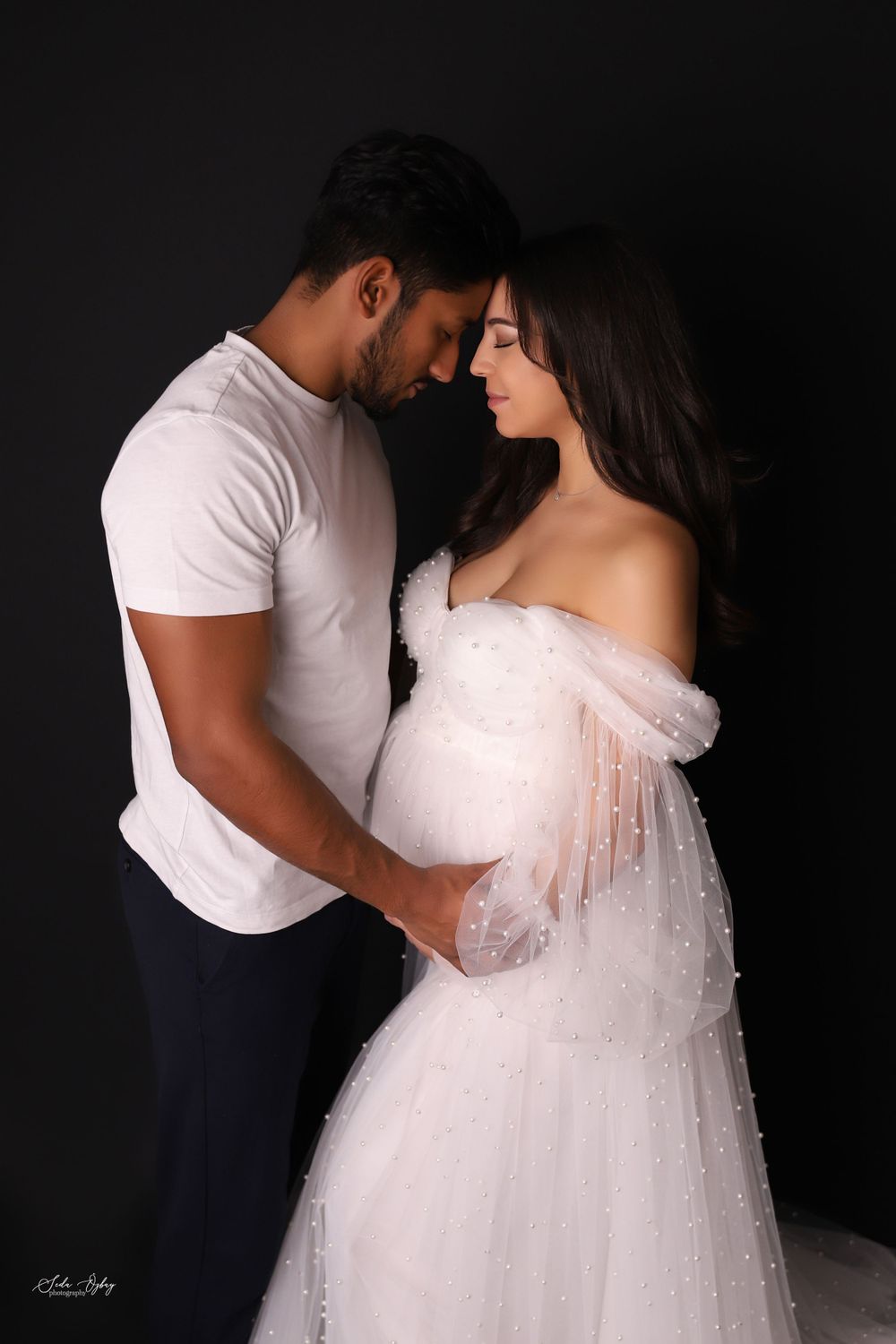 Intimate maternity photoshoot featuring couple in white dress against dark background.