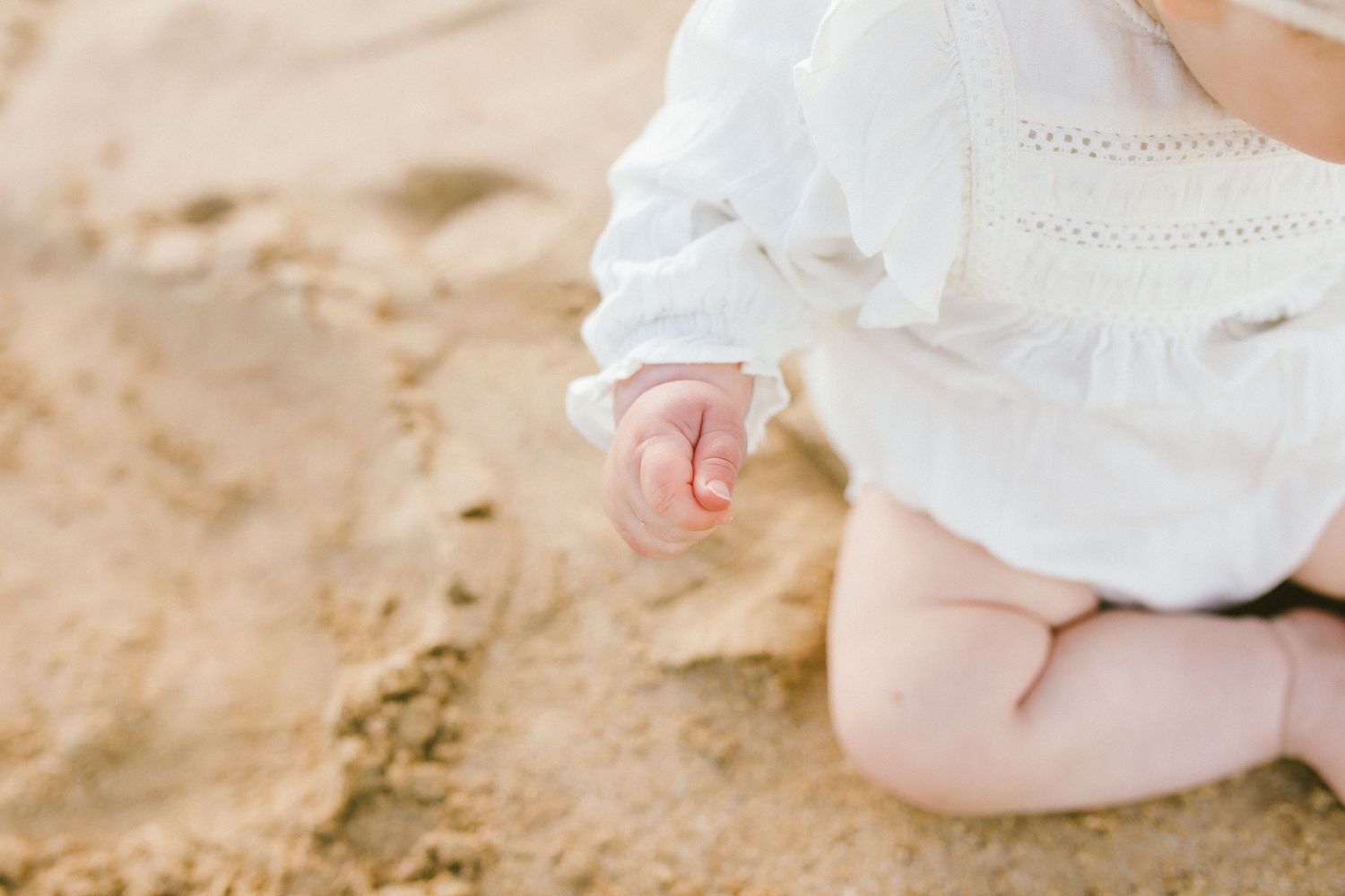 A baby wearing a white dress sits and plays in the sand at the beach.